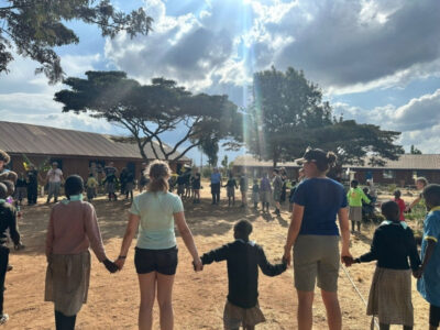 Group of students on a Camps International expedition in Africa at a local school, all standing in a big circle and holding hands with the local children.