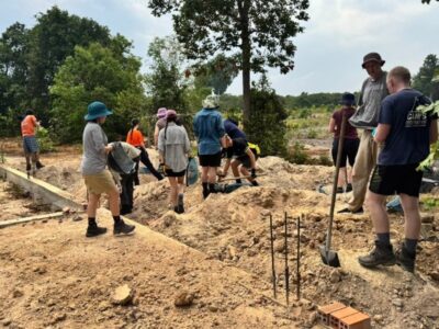 Group of students from Australia on a Camps International expedition in Cambodia working on the foundations for a local Cambodian school. The group is hard at work, some are standing watching with their shovels, waiting to get stuck in, and other students are digging away at the ground. They're all in baggy clothes and bucket hats.