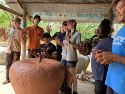 A group of Australian students on a Camps International expedition to Cambodia. They are helping to build clay water storage pots, and are standing around their large clay pot with their hands in the air showing how covered in red clay they are. They're all smiling for the camera and looking off to another photo being taken on the left.