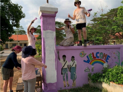 Group of students on a Camps International expedition in Cambodia, stood on a pink wall with a mural. They're painting it and smiling.