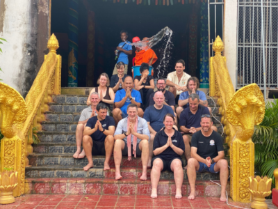 A group of teachers on a familiarisation trip in Cambodia with Camps International all sat on some golden steps being blessed by a local monk, by having water thrown over them.