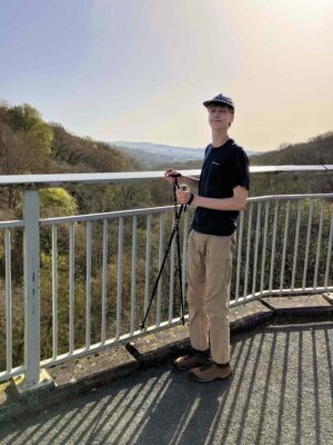 a young boy wearing a hat and a black t-shirt, holding walking poles and smiling with lots of trees behind him