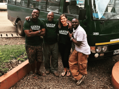 three camps international staff from Tanzania posing with a woman wearing camps t-shirt in front of a big green bus
