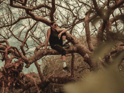 a woman wearing a vest top and leggings, smiling and sitting on a branch on a tree