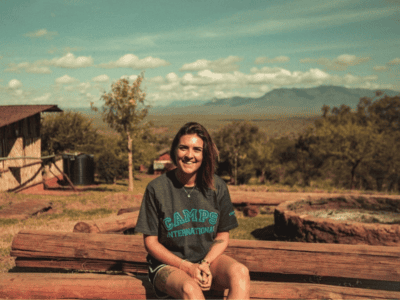 A woman wearing camps international t-shirt smiling and sitting in a bench made from wood in Kenya