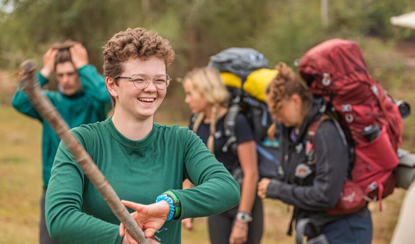 a boy traveller smiling and holding a walking stick with other travellers with backpacks in the background