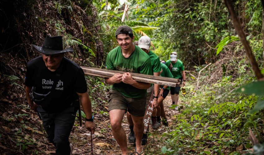 group of traveller students walking in the jungle carrying plank of wood and smiling