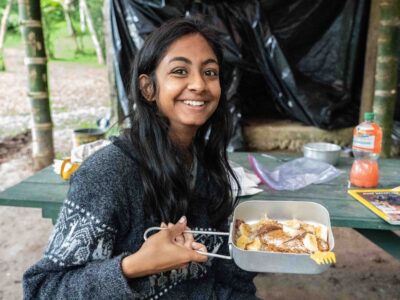 a girl traveller smilling wearing a jumper and holding a cooking pan with a banana chocolate pancake