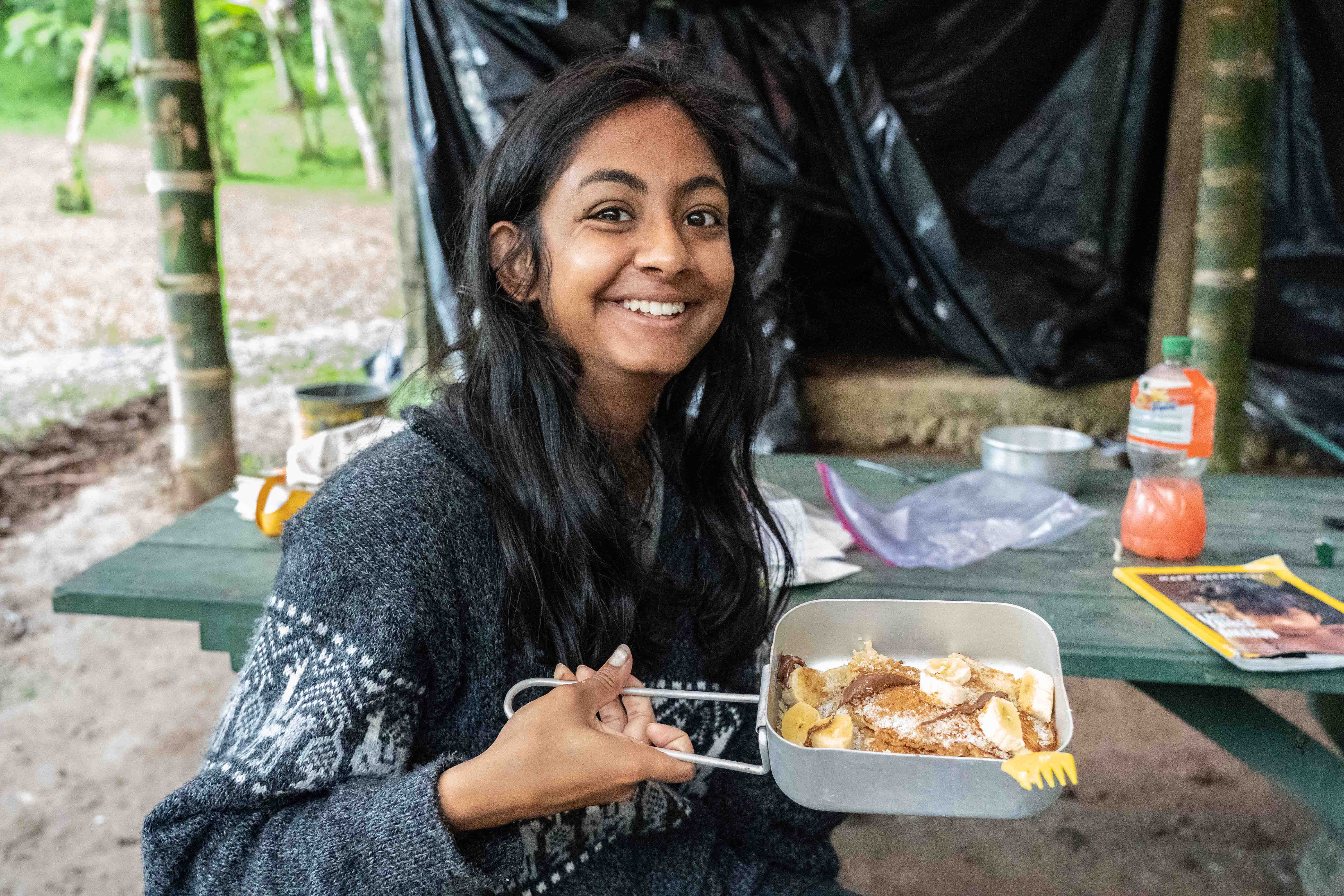 a girl traveller smilling wearing a jumper and holding a cooking pan with a banana chocolate pancake