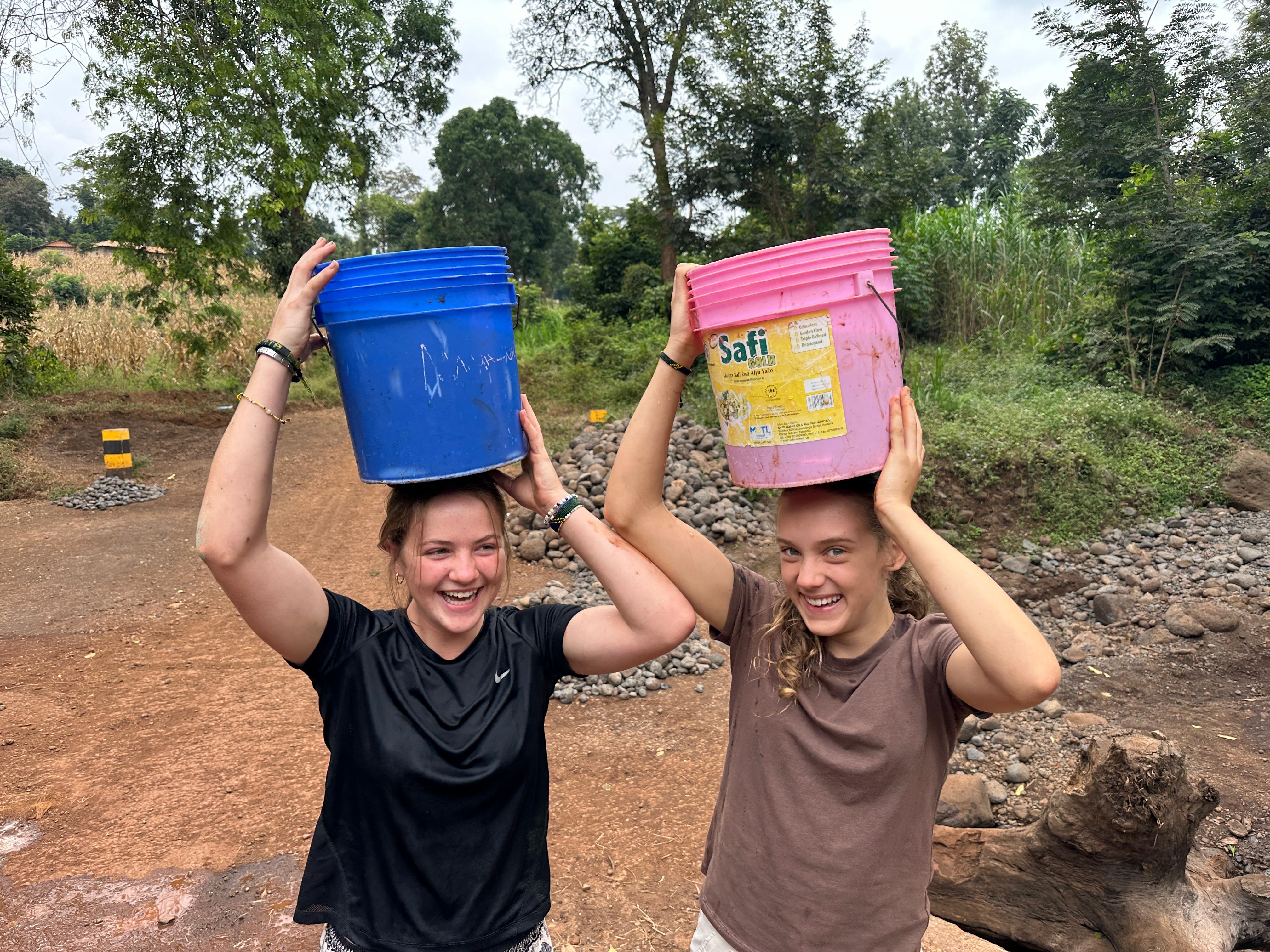 two girls carrying water buckets on their heads and smiling in Tanzania